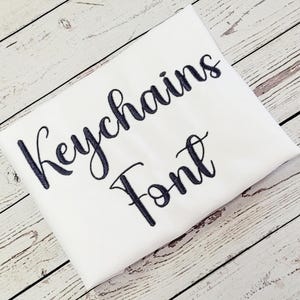 May include: A white fabric item with the words "Keychains Font" embroidered in a dark blue script. The fabric is folded and placed on a light wood-grain surface.