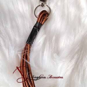 Pode incluir: Um porta-chaves de couro castanho com uma pega entrançada e um anel prateado. O porta-chaves tem um feixe de finas tiras de couro na extremidade, presas com fita adesiva preta. O texto "Seraphinas Accessoires" é visível.