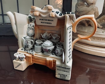May include: A decorative Portmeirion teapot shaped like a shop, with miniature teacups, teapots, and a "Portmeirion Potteries" sign. The teapot has a white and orange handle and floral patterns. The base reads "Fragile" and "Handle with Care."