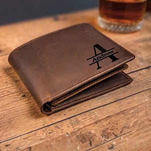May include: A brown leather bifold wallet featuring a dark engraved design on the front. The engraving consists of a large capital letter A with the name Andrew written horizontally across the center of the letter in a clean, sans-serif font.