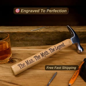 May include: A steel hammer with a light-colored wooden handle engraved with the text The Man. The Myth. The Legend. The hammer is positioned near a glass containing an amber liquid and an orange pencil. The image includes the text Engraved To Perfection.