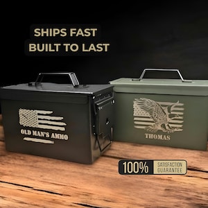 May include: Two metal ammo boxes, one black and one olive green, sit on a wooden surface. The black box features an American flag design and the text "OLD MAN'S AMMO." The green box has an eagle graphic and the name "THOMAS."