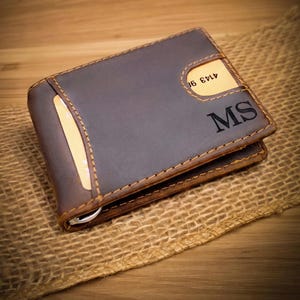May include: A dark brown leather bifold wallet featuring visible tan stitching, a metal money clip, and two card slots. The front of the wallet is embossed with the letters MS and contains a partially visible credit card with the numbers 4143 98.