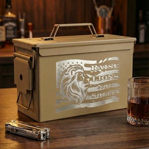 May include: A tan metal ammunition box featuring a laser-engraved design of a lion's head superimposed over a stylized American flag. The text RAISE LIONS NOT SHEEP is engraved to the right of the lion. The box has a top handle and side latch.