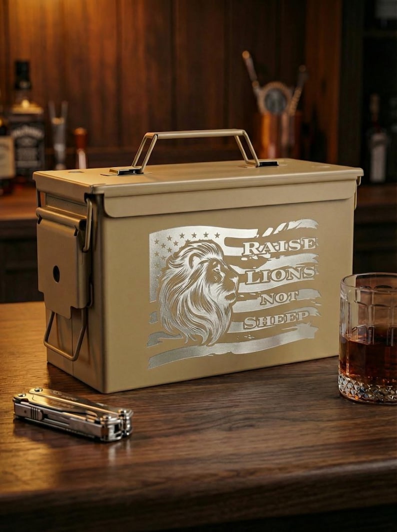 May include: A tan metal ammunition box featuring a laser-engraved design of a lion's head superimposed over a stylized American flag. The text RAISE LIONS NOT SHEEP is engraved to the right of the lion. The box has a top handle and side latch.