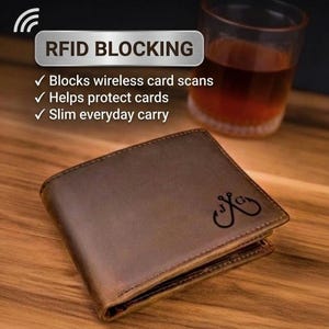 May include: A brown leather bifold wallet featuring a dark engraved logo of two crossed fishing hooks with the letters J and G. Text above reads RFID BLOCKING, Blocks wireless card scans, Helps protect cards, and Slim everyday carry.