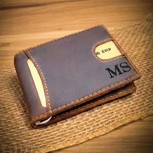 May include: A dark brown leather wallet featuring visible tan stitching, a metal money clip, and two card slots. The initials MS are embossed on the front surface. One card slot contains a light-colored card with visible numbers, and another slot holds a card.