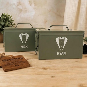 May include: Two olive green metal ammunition boxes with hinged lids and carrying handles. Each box features a white graphic of a tuxedo jacket with a bowtie and the printed names NICK and RYAN. A rectangular wooden plaque reads GROOMSMEN.