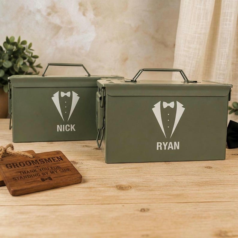 May include: Two olive green metal ammunition boxes with hinged lids and carrying handles. Each box features a white graphic of a tuxedo jacket with a bowtie and the printed names NICK and RYAN. A rectangular wooden plaque reads GROOMSMEN.