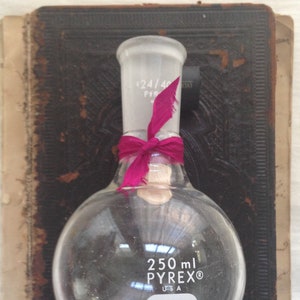 May include: A clear, round-bottomed 250 ml Pyrex flask with a ground glass joint, tied with a magenta ribbon. The flask is labeled with "250 ml PYREX® USA". The flask rests on a vintage book with a dark cover.