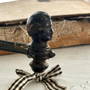 May include: A dark, weathered metal handle featuring a sculpted human head at the end, tied with a small black and white gingham ribbon. The metal shows signs of age with a textured, matte finish and visible oxidation, extending into a straight bar.