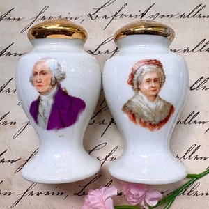 May include: A pair of white ceramic salt and pepper shakers with gold-colored lids. Each shaker features a portrait of a historical figure. One portrait is of a man in a purple coat, and the other is of a woman in a red hat. Pink flowers are in the foreground.