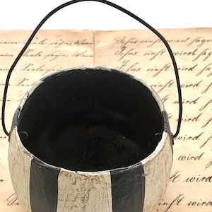 May include: A small, round, hand-painted container with a black interior and a black and white striped exterior. It has a black wire handle. The container sits on a vintage paper background.