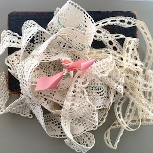 May include: A collection of ivory-colored lace ribbons with various openwork patterns. A small pink paper bow is nestled among the ribbons. The lace is arranged on a dark, textured surface, creating a vintage aesthetic.