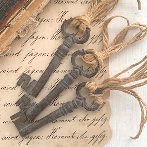 May include: Three antique-style metal keys with decorative handles, tied with twine. The keys rest on a vintage paper with handwritten script. The paper has a distressed, aged appearance, and the keys have a rustic, weathered finish.