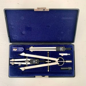 May include: A set of silver and black drafting compasses and tools in a dark blue case. The case is open, revealing the tools neatly arranged on a blue velvet-like surface. A small pencil sharpener is also included.