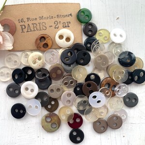 May include: A collection of round, two-hole buttons in various colours including black, white, brown, clear, and green. A tan rectangular tag is present with the printed text 16, Rue Marie-Stuart PARIS - 2e ar, alongside a pale pink fabric flower.