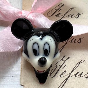 K&ouml;nnte beinhalten: Eine Mickey Mouse-Kopf-Figur in Schwarz, Wei&szlig; und Rot. Die Figur hat gro&szlig;e schwarze Ohren, ein wei&szlig;es Gesicht und einen roten Mund. Im Hintergrund ist ein rosa Band zu sehen.