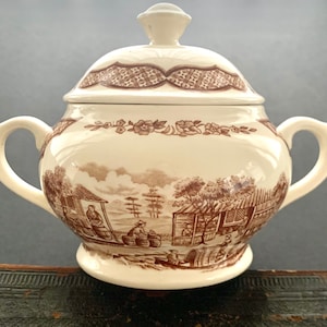 May include: Cream-colored ceramic sugar bowl with a lid and two handles. The bowl features a brown transferware design depicting a rural scene. The lid has a decorative pattern and a small knob.