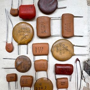 May include: A collection of various electronic capacitors in shades of brown, orange, and red. These components feature metallic leads and printed technical specifications, including alphanumeric codes like RMC 3KV280, 104M, and 220J, arranged in a scattered layout.