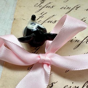 K&ouml;nnte beinhalten: Eine schwarz-wei&szlig;e Keramik-Vogelfigur liegt auf einer hellrosa Satin-Schleife. Die Schleife ist gebunden und liegt auf einem Vintage-Papierhintergrund mit handschriftlichem Schriftzug. Der Vogel hat ein gl&auml;nzendes Finish.