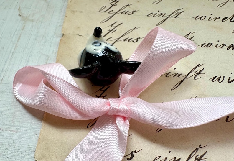 K&ouml;nnte beinhalten: Eine schwarz-wei&szlig;e Keramik-Vogelfigur liegt auf einer hellrosa Satin-Schleife. Die Schleife ist gebunden und liegt auf einem Vintage-Papierhintergrund mit handschriftlichem Schriftzug. Der Vogel hat ein gl&auml;nzendes Finish.