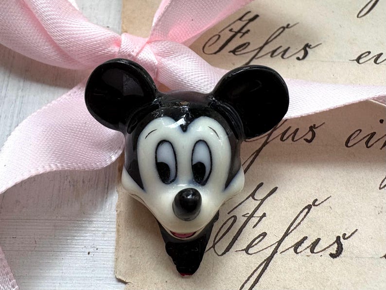 K&ouml;nnte beinhalten: Eine Mickey Mouse-Kopf-Figur in Schwarz, Wei&szlig; und Rot. Die Figur hat gro&szlig;e schwarze Ohren, ein wei&szlig;es Gesicht und einen roten Mund. Im Hintergrund befindet sich ein rosa Band.
