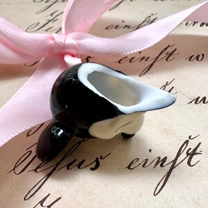K&ouml;nnte beinhalten: Kleine, schwarz-wei&szlig;e Keramik-Orca-Wal-Figur mit wei&szlig;em Innenraum. Die Figur befindet sich auf einem Vintage-Papierhintergrund mit Kursivschrift und einem rosa Band.