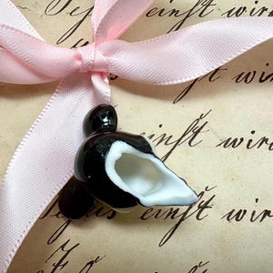 K&ouml;nnte beinhalten: Eine schwarz-wei&szlig;e Keramik-Vogelfigur mit wei&szlig;em Innenraum, gebunden mit einem hellrosa Satinband. Der Vogel sitzt auf einem Vintage-Papier mit handschriftlichem Schriftzug. Das Band ist zu einer Schleife gebunden.
