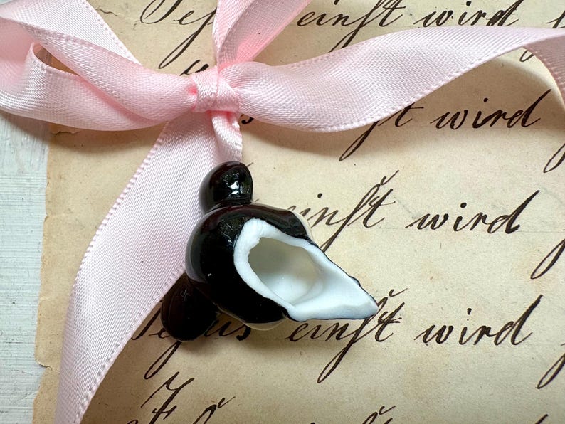 K&ouml;nnte beinhalten: Eine schwarz-wei&szlig;e Keramik-Vogelfigur mit wei&szlig;em Innenraum, gebunden mit einem hellrosa Satinband. Der Vogel sitzt auf einem Vintage-Papier mit handschriftlichem Schriftzug. Das Band ist zu einer Schleife gebunden.