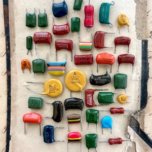May include: A collection of various electronic capacitors in assorted shapes, sizes, and colours including red, green, yellow, blue, orange, and black. Each component features two metal wire leads extending from the body, with some displaying printed technical specifications and values.