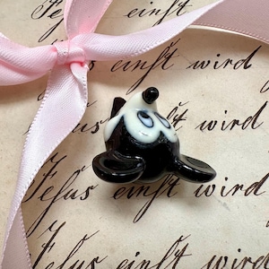 K&ouml;nnte beinhalten: Eine kleine Robbenfigur aus Glas in Schwarz und Wei&szlig; mit einer rosa Schleife. Die Robbe hat eine gl&auml;nzende Oberfl&auml;che und befindet sich auf einem Vintage-Papierhintergrund mit handschriftlichem Text.