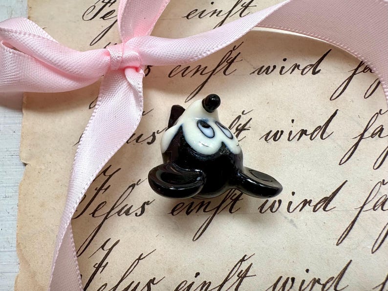 K&ouml;nnte beinhalten: Eine kleine Robbenfigur aus Glas in Schwarz und Wei&szlig; mit einer rosa Schleife. Die Robbe hat eine gl&auml;nzende Oberfl&auml;che und befindet sich auf einem Vintage-Papierhintergrund mit handschriftlichem Text.