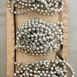 May include: Three separate strands of metallic silver beads arranged in coiled loops on a rectangular piece of aged, tan-colored paper featuring cursive script text. The beads have a reflective, shiny finish and are strung together in a continuous line.