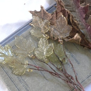 May include: A collection of translucent pale yellow glass maple leaf ornaments, each attached to a thin, dark brown wire stem. The leaves feature detailed veining and are arranged in a cluster, showcasing a clear, textured surface and a delicate, botanical-inspired design.