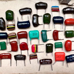 May include: A collection of various electronic capacitors in multiple shapes and colors, including red, green, blue, yellow, brown, and black. Each component features two metal leads and printed technical specifications on the surface.