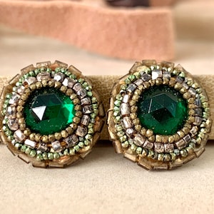 May include: Two round earrings with a central, faceted emerald green stone. The earrings are intricately beaded with gold, silver, and green beads. The design is set against a neutral background.