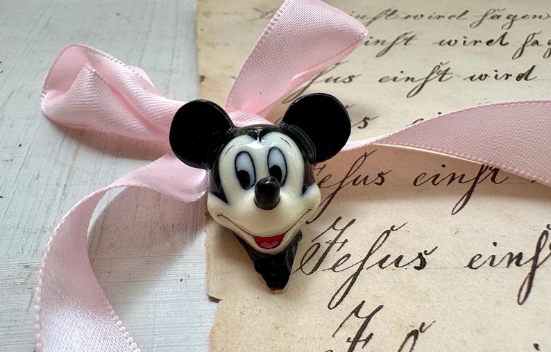 K&ouml;nnte beinhalten: Eine Mickey Mouse Kopf-Figur mit schwarzen Ohren, wei&szlig;em Gesicht und rotem Mund. Ein hellrosa Band ist um die Figur geschlungen. Der Hintergrund zeigt Vintage-Papier mit handschriftlichem Skript.