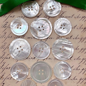May include: A collection of iridescent, pearlescent buttons with four holes, arranged on a vintage paper background. A green ribbon is tied at the top. The buttons vary in size and have a subtle, swirling pattern.