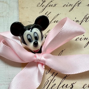 K&ouml;nnte beinhalten: Eine Mickey Mouse-Figur befindet sich auf einer hellrosa Satin-Schleife. Die Figur ist schwarz, wei&szlig; und rot. Der Hintergrund ist ein Vintage-Papier mit handschriftlichem Skript.