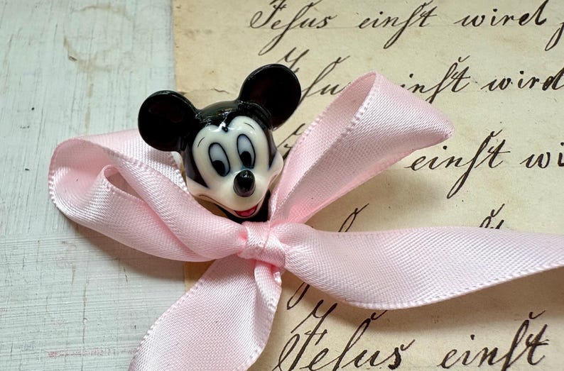K&ouml;nnte beinhalten: Eine Mickey Mouse-Figur befindet sich auf einer hellrosa Satin-Schleife. Die Figur ist schwarz, wei&szlig; und rot. Der Hintergrund ist ein Vintage-Papier mit handschriftlichem Skript.