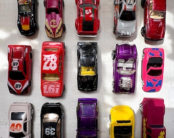 May include: A collection of toy race cars in various colors, including red, black, purple, and yellow. Some cars feature racing numbers and graphics. The cars are arranged on a light-colored surface, showcasing their diverse designs.