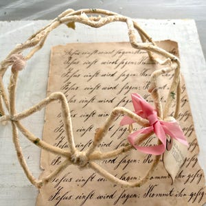 May include: A vintage-style, beige-colored decorative crown made of twisted material, adorned with a pink ribbon and a small tag. The crown rests on a piece of aged paper with handwritten script.