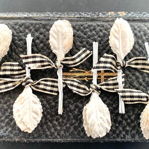 May include: Six off-white artificial leaf sprigs attached to thin white wire stems, each tied with a black and white gingham ribbon. The sprigs are arranged in two rows, with three sprigs pointing upward and three sprigs pointing downward.