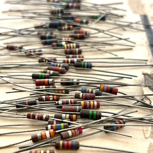 May include: A collection of numerous cylindrical electronic resistors with various coloured bands indicating resistance values. Each resistor features two long, straight metal wire leads extending from opposite ends of the small, dark-coloured body.