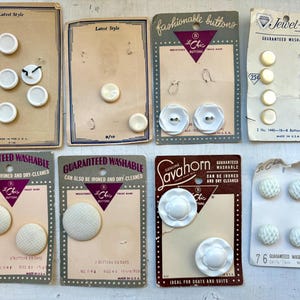 May include: A selection of vintage white buttons on their original cards. Styles include round, flower-shaped, and textured designs. Some cards display the text "Latest Style" and "Guaranteed Washable". Measurements are in inches.
