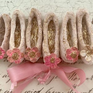 Peut inclure: Six chaussons de danse miniatures de couleur crème pâle avec des accents de paillettes dorées à l'intérieur et à l'extérieur. Chaque chausson présente une petite fleur rose ou crème au centre doré pailleté, liés par un ruban rose.