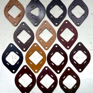 May include: A collection of fourteen diamond-shaped leather patches in various shades of brown, tan, black, and maroon. Each patch features a central square cutout, four small rectangular slots, and two circular holes at the top and bottom points.
