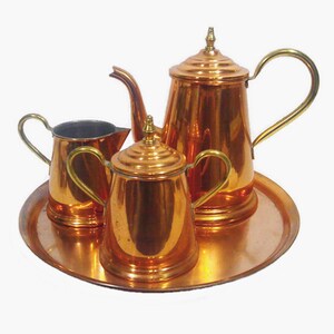 May include: A vintage copper tea set with a teapot, sugar bowl, creamer, and serving tray. The set has a shiny, reflective surface and gold-colored handles. The teapot is approximately 8 inches tall.