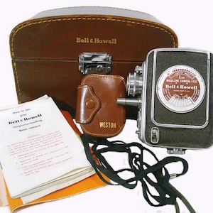 May include: A vintage Bell & Howell 8mm magazine camera with a brown leather case and a Weston leather accessory case. The camera has a black body with a silver lens and a dial. A manual and a black strap are also included.